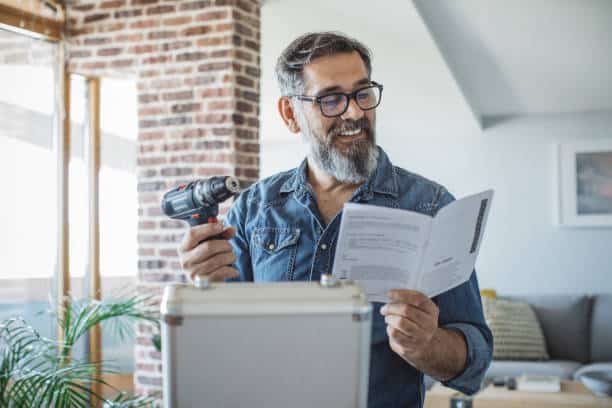A man with glasses and a beard is holding a power drill and reading an instruction manual. He is standing in a room with a brick wall.