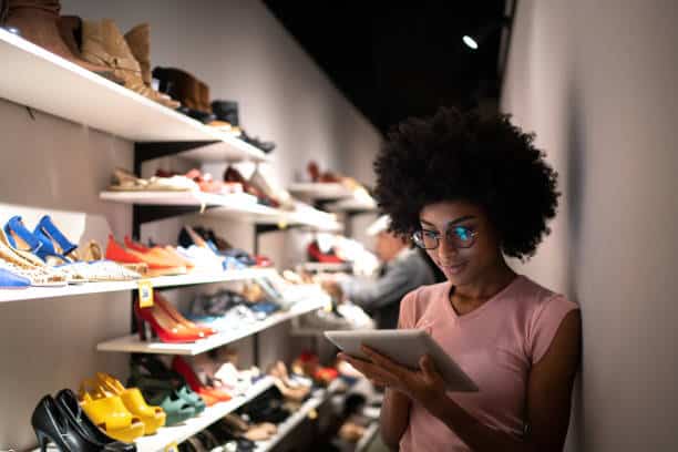 A person with curly hair and glasses stands in a shoe store, holding a tablet while other shoppers browse shoes on shelves in the background.