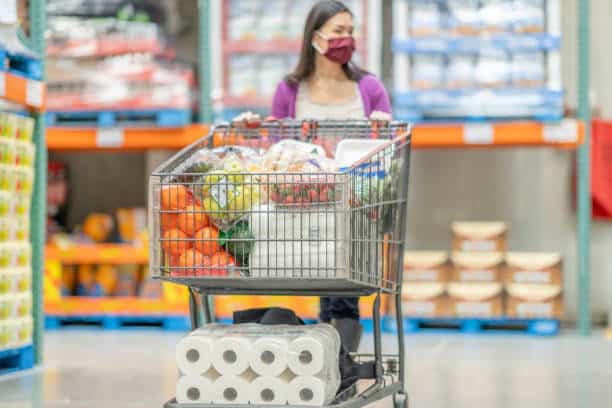 A woman wearing a mask pushes a shopping cart filled with groceries, including fruits, vegetables, and paper towels, in a store.