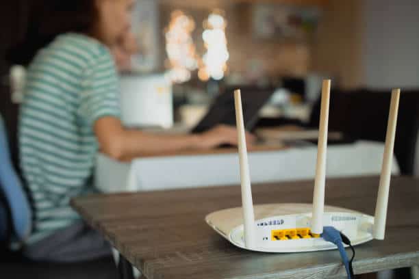 A person works on a laptop at a table in the background while a Wi-Fi router with three antennas sits on a nearby surface in the foreground.