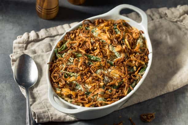A white casserole dish filled with a green bean casserole topped with crispy fried onions, placed on a beige cloth next to a spoon.