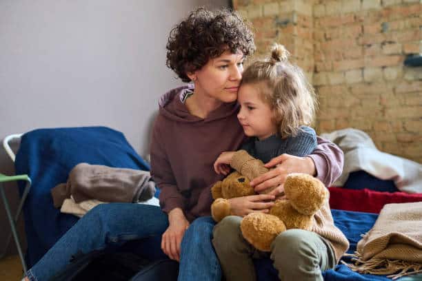 A woman with curly hair sits on a bed holding a young child who is hugging a teddy bear. They are in a room with exposed brick walls and folded clothes nearby.