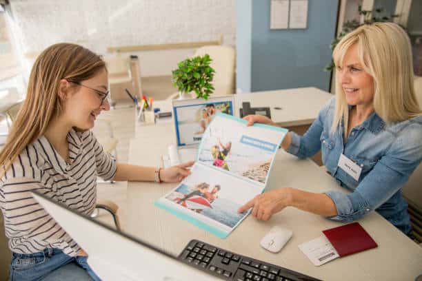 Two women are sitting at a desk in an office. One is showing the other a brochure with travel photos. A computer screen and various office supplies are on the desk.