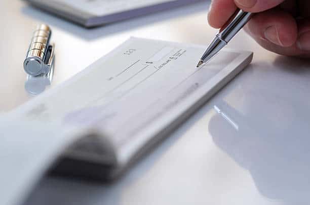 A hand holding a pen writes on a check, with a metallic pen cap placed on the white surface nearby.