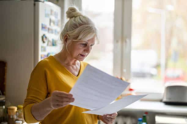An elderly woman with gray hair in a bun reads documents in a kitchen, standing near a refrigerator.