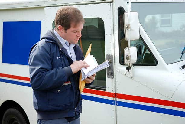 Postal worker in a blue uniform sorting mail and envelopes beside a white mail truck with a blue and red stripe.
