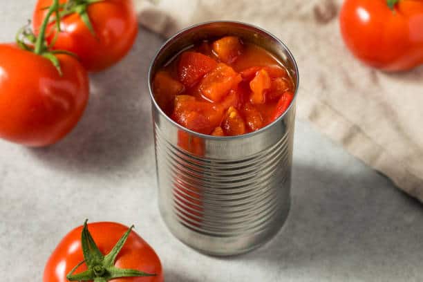 A can of diced tomatoes sits on a counter with three fresh tomatoes nearby and a beige cloth in the background.
