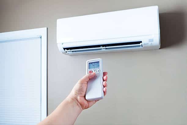 A hand holds a remote control, pointing it at a wall-mounted air conditioning unit.