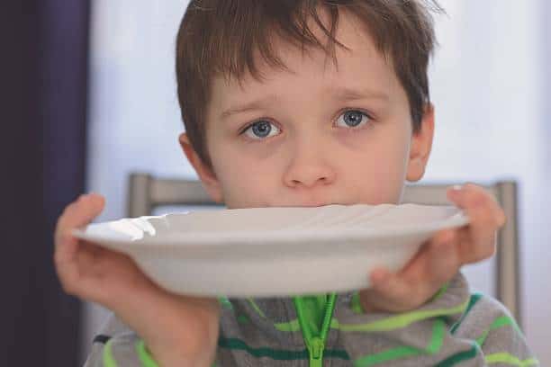 A young boy holds an empty white bowl up to his mouth, gazing directly at the camera. He is wearing a gray and green striped shirt.