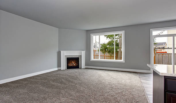 A spacious empty living room with light grey walls, a large window, a white fireplace, and grey carpet flooring. Natural light enters through the window, and the kitchen is partially visible.