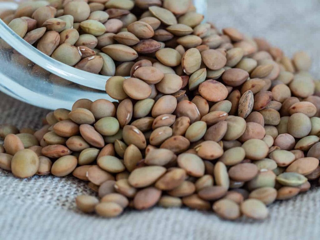 A glass bowl tipped over, spilling brown lentils onto a textured beige surface.