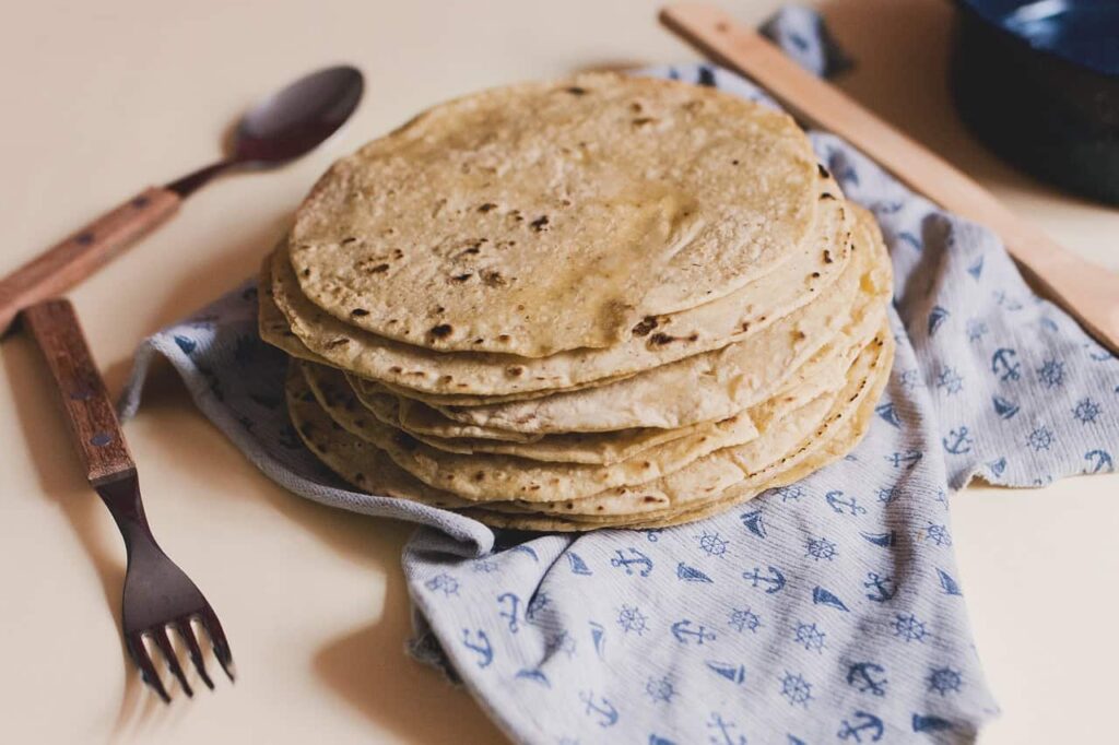 A stack of homemade tortillas placed on a blue-patterned cloth, with a wooden-handled spoon and fork next to it on a beige surface.