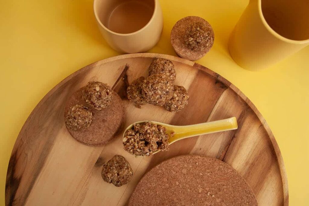 A wooden tray holds granola clusters, a spoon with some clusters, and two cups on a yellow surface.