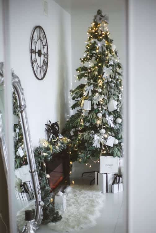 A decorated Christmas tree with white ornaments and lights stands beside a fireplace, a large mirror, and various gift boxes in a cozy, white room.