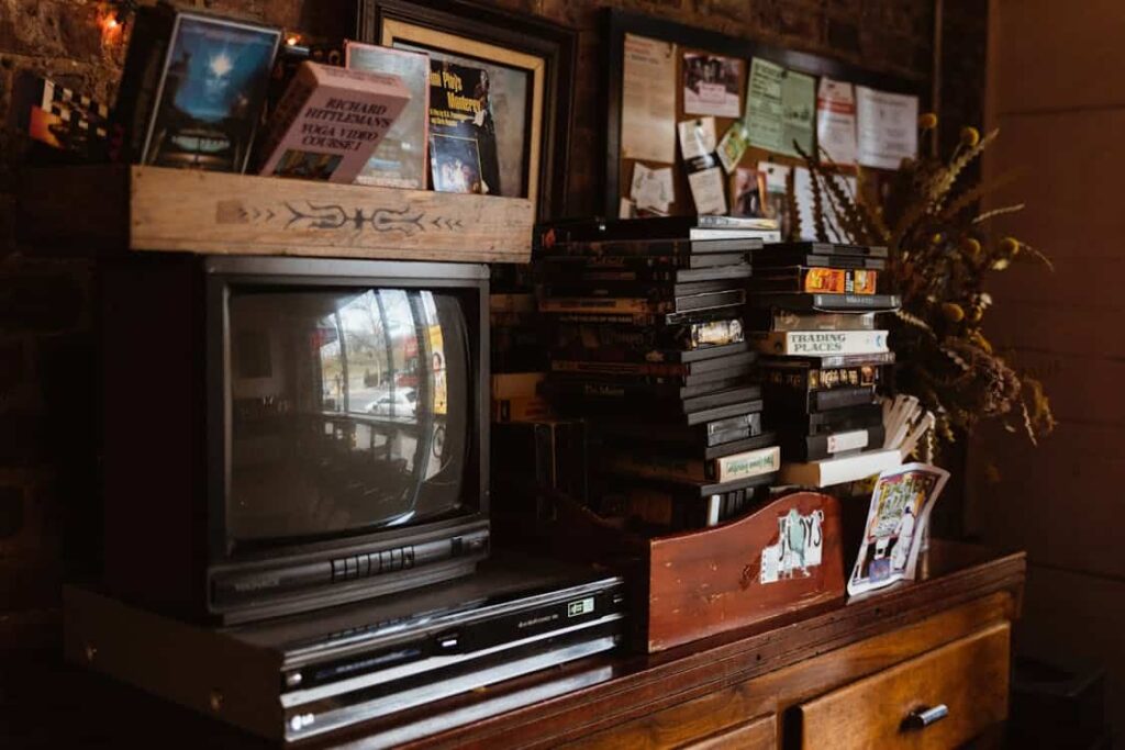 A vintage television and VCR sit on a wooden dresser, surrounded by VHS tapes, books, and a collection of framed photos and papers pinned to a bulletin board.