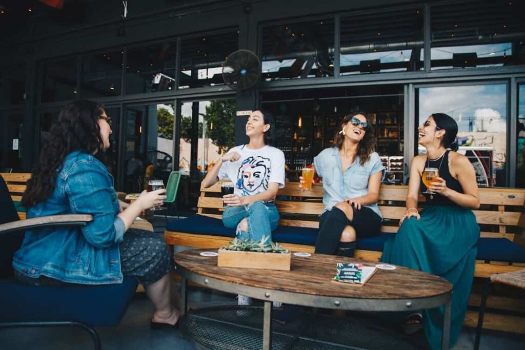 Four people are sitting outdoors at a wooden table, laughing and enjoying drinks at a bar or café, discussing how life priorities shift with older age.