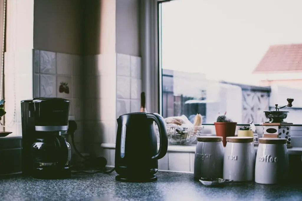 Kitchen countertop with a coffee maker, electric kettle, and various jars and containers next to a window.