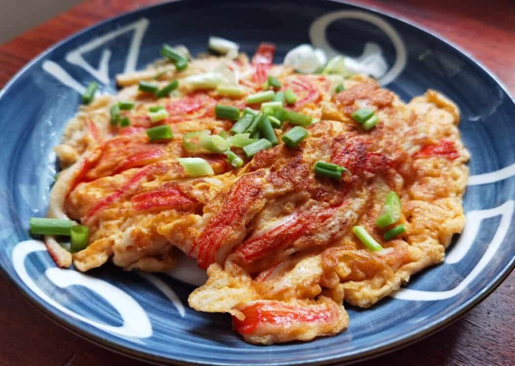 A blue plate with an omelet topped with green onions and a red ingredient, possibly crab sticks, set on a wooden surface.