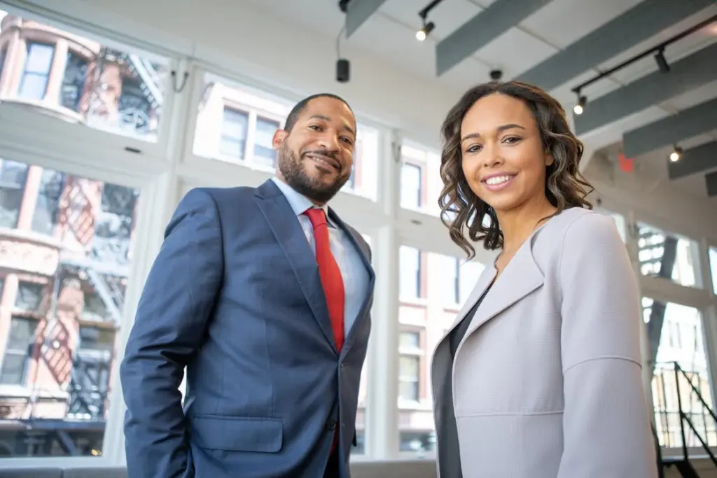 Two professionally dressed individuals, a man in a blue suit with a red tie, and a woman in a light grey blazer, standing and smiling in a brightly lit office space with large windows.