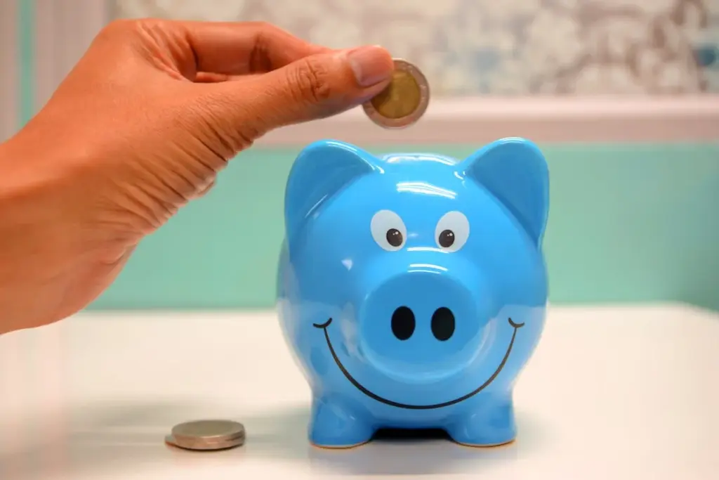 Hand depositing a coin into a smiling blue piggy bank, with another coin placed on the table beside it.