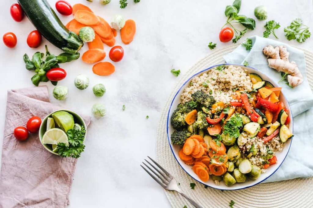A bowl of mixed vegetables and quinoa on a white table, surrounded by scattered vegetables, lime slices, and a fork.