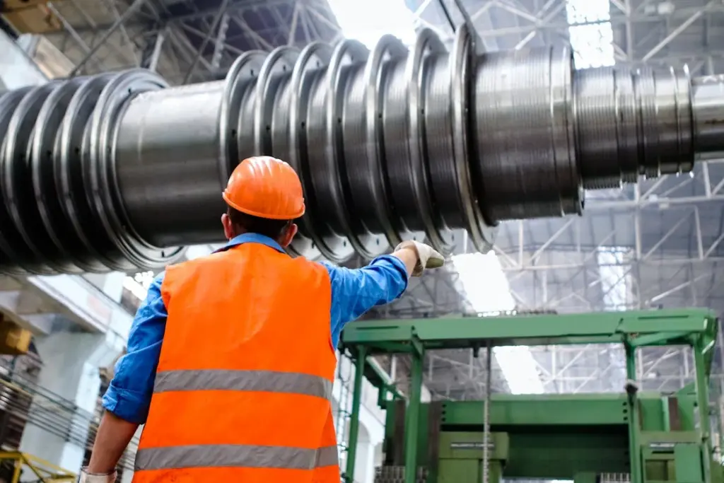 A worker in an orange safety vest and helmet inspects a large industrial machine with multiple coils in a factory setting.