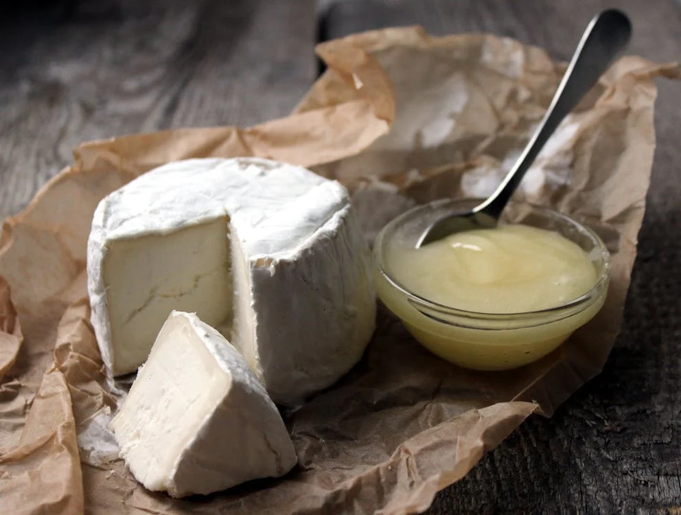Round of soft cheese with a wedge cut out, placed on brown parchment paper, next to a small bowl of honey with a spoon on a rustic wooden surface.