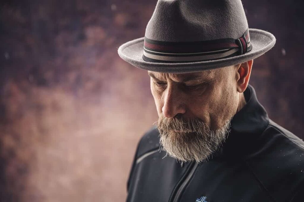 A man with a grey beard wearing a grey fedora and black jacket looks down, set against a blurred, textured background.