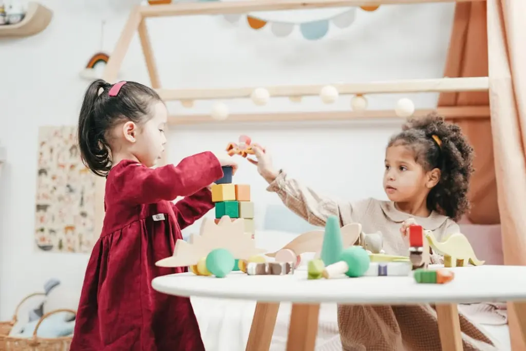 Two young girls are playing with colorful building blocks and toy figures at a round table in a well-lit room.