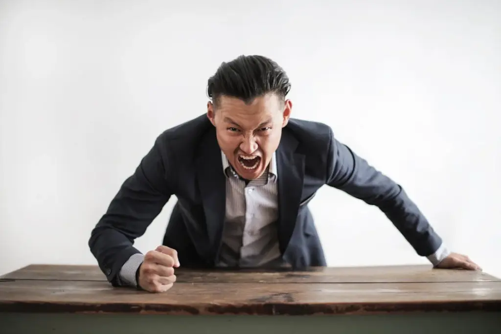 A man in a suit leans forward on a table, yelling with his fist clenched.