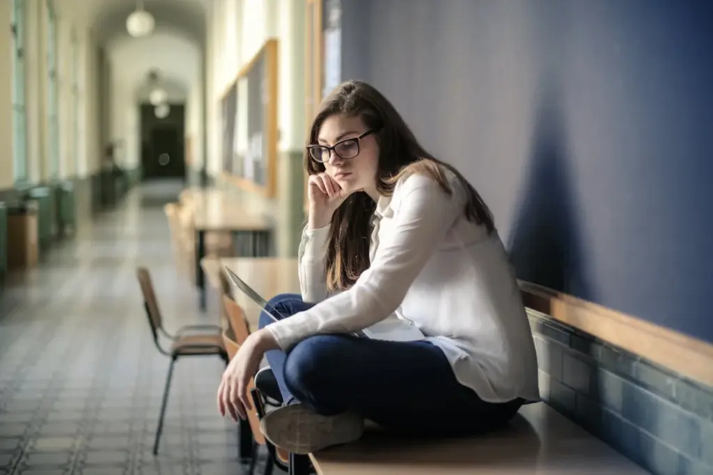 A woman with glasses, wearing a white shirt and blue jeans, sits cross-legged on a desk in a corridor with a contemplative expression.