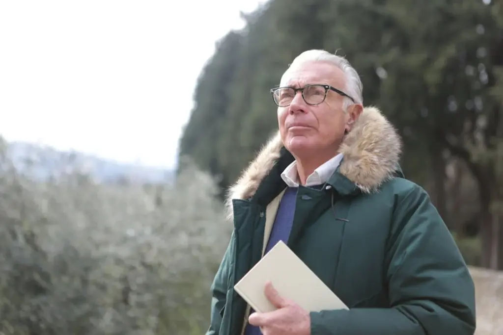 Elderly man with glasses and a green coat holding a book, standing outdoors near trees.