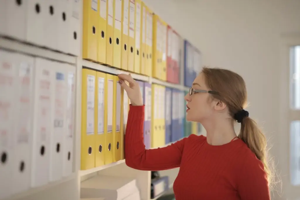 A woman in a red sweater and glasses is reaching for a yellow binder from a shelf filled with various colored binders in an office setting.