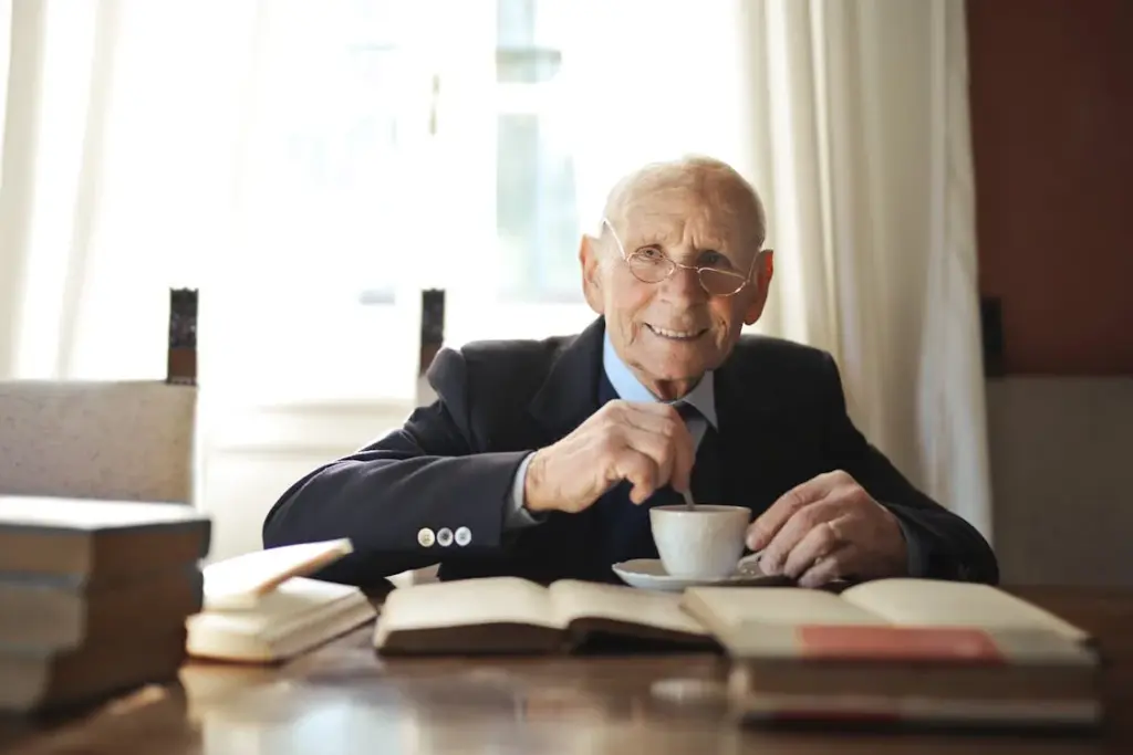 Elderly man in a suit and glasses sits at a table, smiling and stirring a cup of coffee, with books open in front of him.