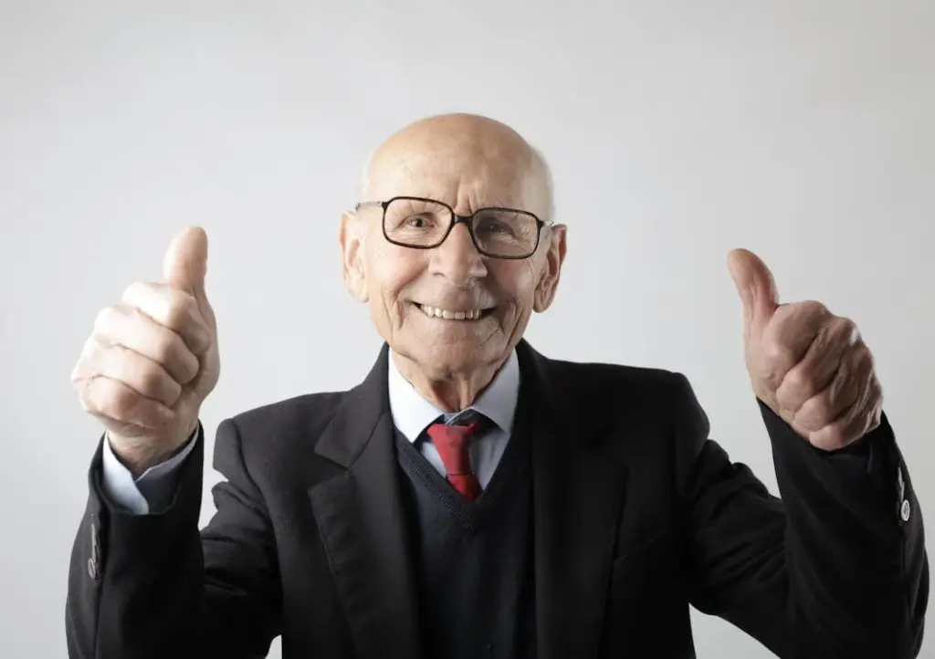 Elderly man in a suit and glasses smiling and giving a thumbs up gesture with both hands against a plain background.