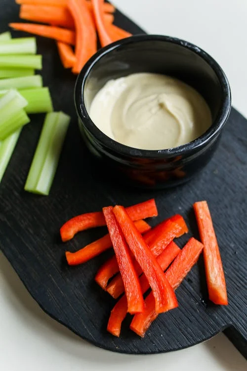 A black tray with sliced celery and red bell peppers surrounding a black bowl of creamy dip.