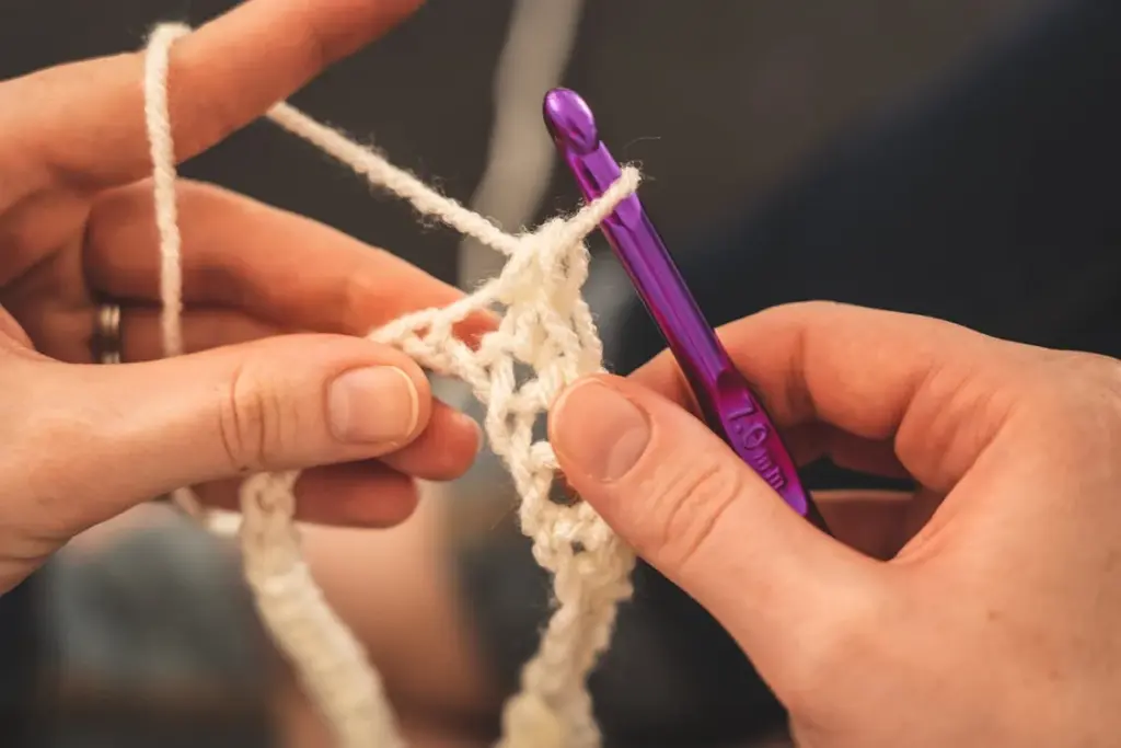 Close-up of hands crocheting with a purple crochet hook and white yarn.