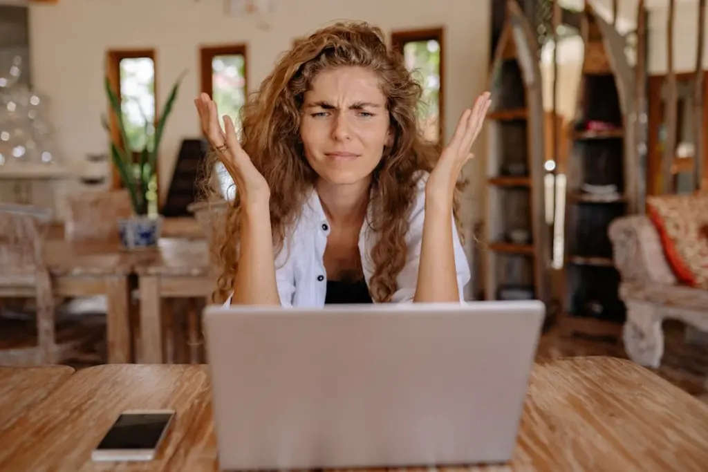 A person with curly hair sits at a wooden table in front of a laptop, looking frustrated with both hands raised. A smartphone is placed on the table beside them.