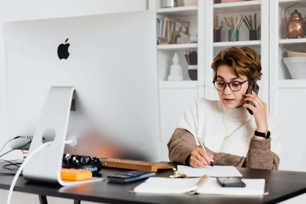 A person with short hair wearing glasses and a sweater is sitting at a desk with two monitors, talking on a phone and writing notes. The desk has various items, including a notebook and a smartphone.