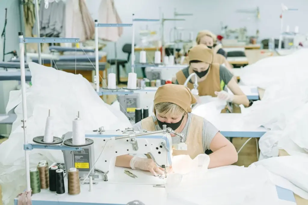 Workers wearing headscarves and masks sit at sewing machines, assembling fabric in a well-lit room filled with sewing equipment and materials.
