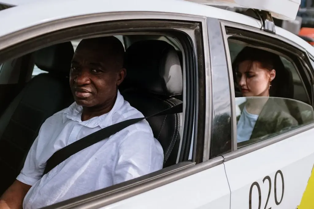 A man in a white shirt drives a taxi while a woman sits in the back seat, looking down, with a seatbelt fastened.