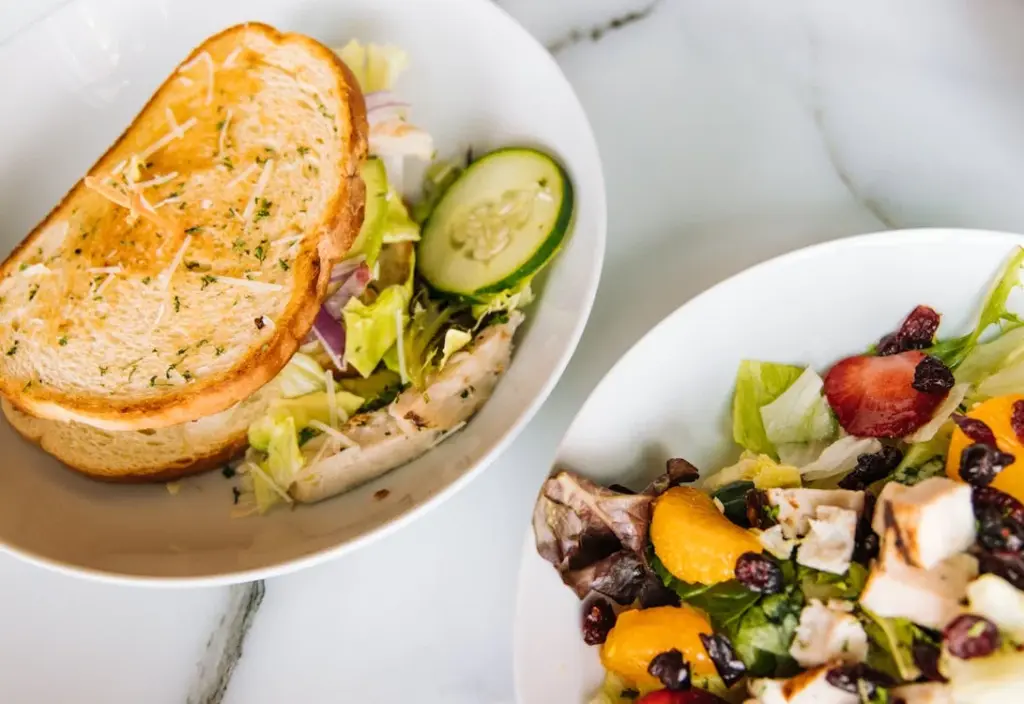 Two bowls of food: one contains a sandwich with salad, the other holds a mixed salad with fruits, chicken, and greens, both on a marble surface.