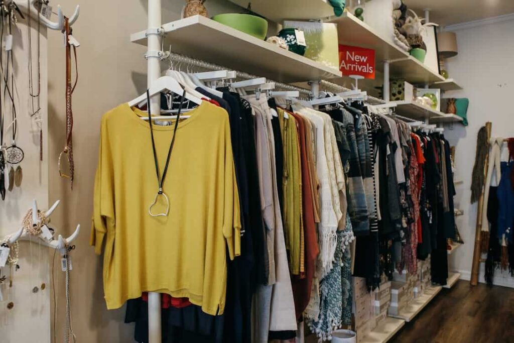 A clothing rack in a boutique displays various tops and jackets, with a yellow long-sleeve shirt and black necklace in the foreground. Shelves above hold accessories and a "New Arrivals" sign.