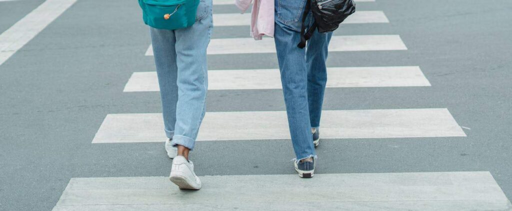 Two people wearing jeans and backpacks walk across a pedestrian crosswalk on a paved street.