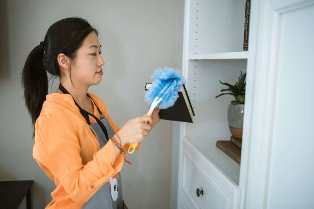 A woman in an orange shirt and gray apron dusts a bookshelf with a blue duster while holding a book.