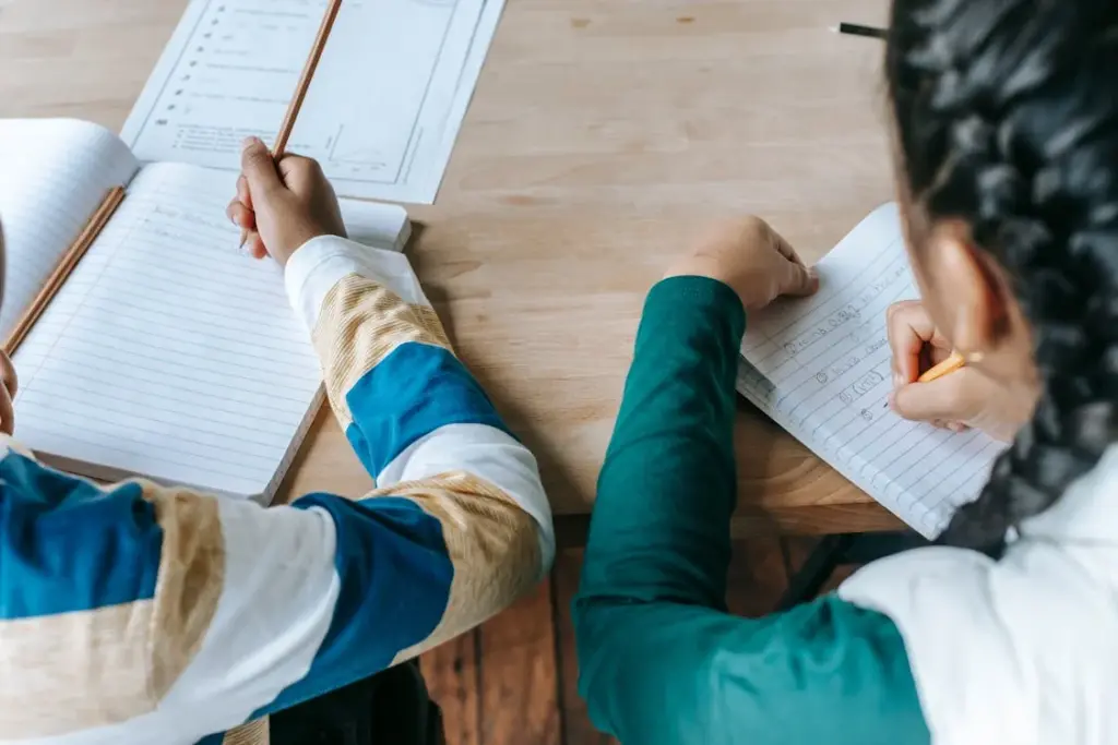Two children sitting at a wooden table, writing in notebooks with pencils. One has a white and blue striped sweater, and the other wears a green sweater. Papers with printed text are also visible.