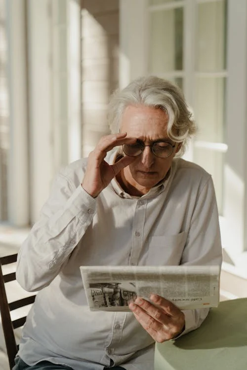 An older man with white hair and glasses reads a newspaper while adjusting his glasses with his left hand. He is sitting indoors near a window.