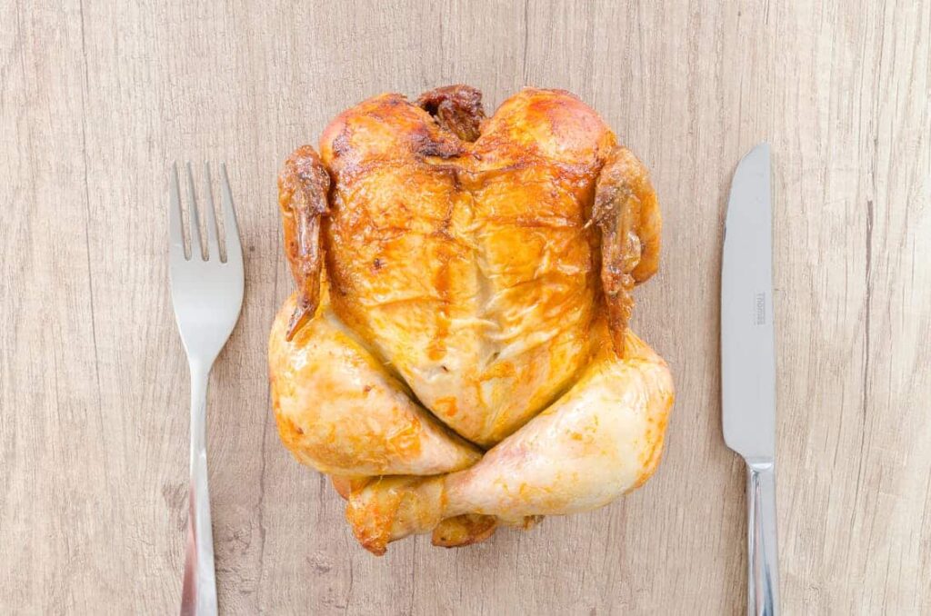 A whole roasted chicken placed on a wooden surface, flanked by a fork on the left and a knife on the right.