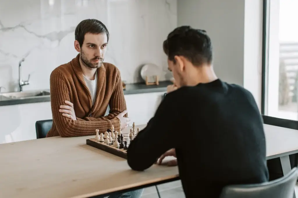 Two men play chess at a table; one, in a brown sweater, watches attentively with folded arms while the other, in a black sweater, contemplates his next move. As they ponder each strategy, it becomes clear that things less important as you get older slip away in the face of this timeless game.