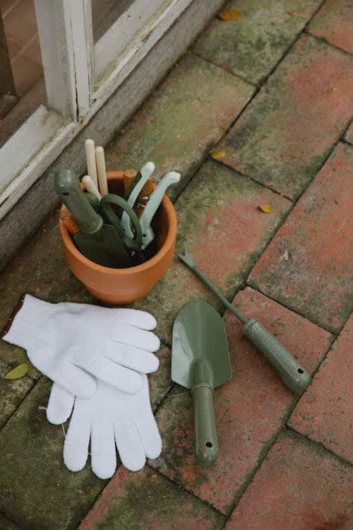 A set of small gardening tools and white gloves are arranged on a brick pavement. The tools include trowels and small shovels, some of which are placed in a terracotta pot.